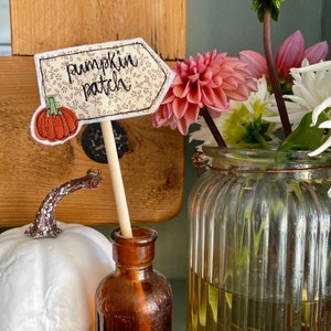 May include: A small, white pumpkin sits next to a brown glass bottle with a wooden stick and a fabric sign that says "pumpkin patch" with a small orange pumpkin on it. The sign is decorated with a floral pattern.