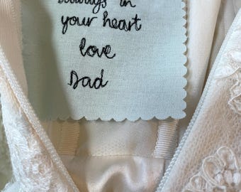 May include: Ivory wedding dress with lace detailing and a light blue fabric tag pinned to the bodice. The tag has embroidered text that reads "always in your heart love Dad". A heart-shaped pin secures the tag.