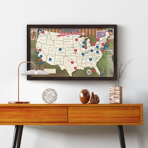 May include: A framed map of the United States with red and blue dots marking locations. The map is titled "Ballpark Travel Quest" and features a baseball theme. The map is hanging on a white wall above a wooden table with decorative objects.