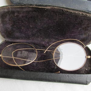 May include: Antique gold-tone eyeglasses with round lenses, resting inside a black leather case with a dark velvet lining. The glasses feature a delicate wire frame and are open, showcasing the lenses.