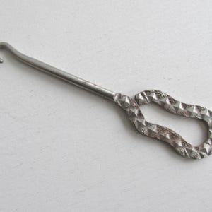 May include: A vintage metal crochet hook with a curved hook end and a textured handle. The handle has a diamond pattern. The hook is silver in color and appears to be made of steel.