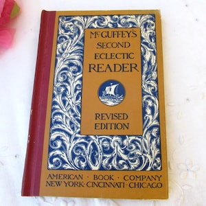Może przedstawiać: Zabytkowa książka zatytułowana "McGuffey's Second Eclectic Reader, Revised Edition". Okładka jest w kolorze brązowym z niebiesko-białym kwiatowym wzorem. Grzbiet jest czerwony. Książka została opublikowana przez American Book Company, z siedzibami w Nowym Jorku, Cincinnati i Chicago.
