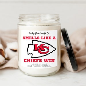 May include: A white soy candle in a glass jar with a red and white label. The label reads "Lucky You Candle Co. Smells Like A Chiefs Win 8 oz/227g Soy Candle Hand-Poured in Telford, PA". The label also features a red and white Kansas City Chiefs logo.