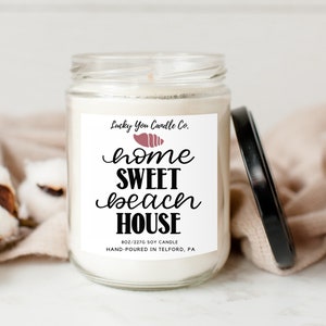 May include: A white soy candle in a glass jar with a black lid. The label is white with black text that reads "Lucky You Candle Co. home SWEET beach HOUSE 8oz/227g Soy Candle Hand-Poured in Telford, PA". There is a pink seashell graphic on the label.