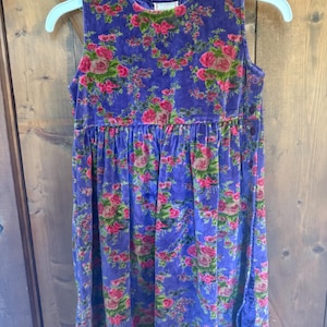 May include: A sleeveless, vintage-style dress in a vibrant purple hue, adorned with a floral pattern of pink and red roses. The dress features a gathered waist and button details, hanging on a light blue hanger. The fabric appears to be a soft velvet.