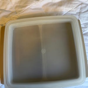 May include: A beige square Tupperware container with a clear lid. The lid has a raised edge and a small circular indentation in the centre. The container is designed for food storage.