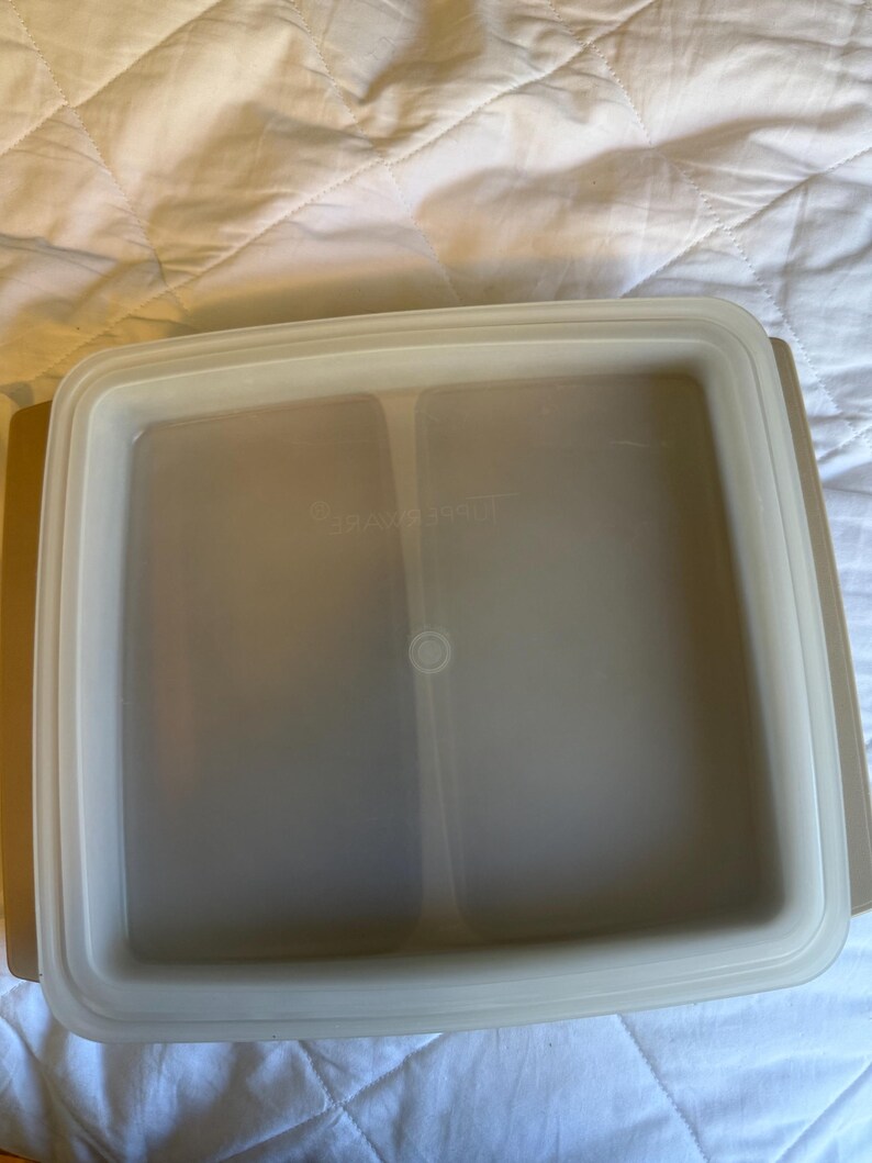 May include: A beige square Tupperware container with a clear lid. The lid has a raised edge and a small circular indentation in the centre. The container is designed for food storage.