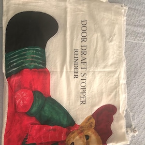 May include: White fabric door draft stopper with a reindeer design. The reindeer is red, green, and black with a brown nose. The text "DOOR DRAFT STOPPER REINDEER" is printed on the fabric.