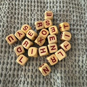 Pode incluir: Um conjunto de dados de letras de madeira com letras vermelhas. Os dados estão espalhados sobre uma superfície texturizada azul clara. Os dados são de cor bege claro.