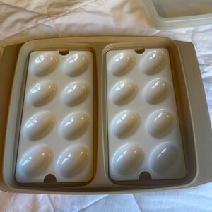 May include: A beige plastic egg container with two trays, each holding six white egg-shaped moulds. The container has a matching lid, designed for storing and transporting eggs. The trays are set inside a larger rectangular base.