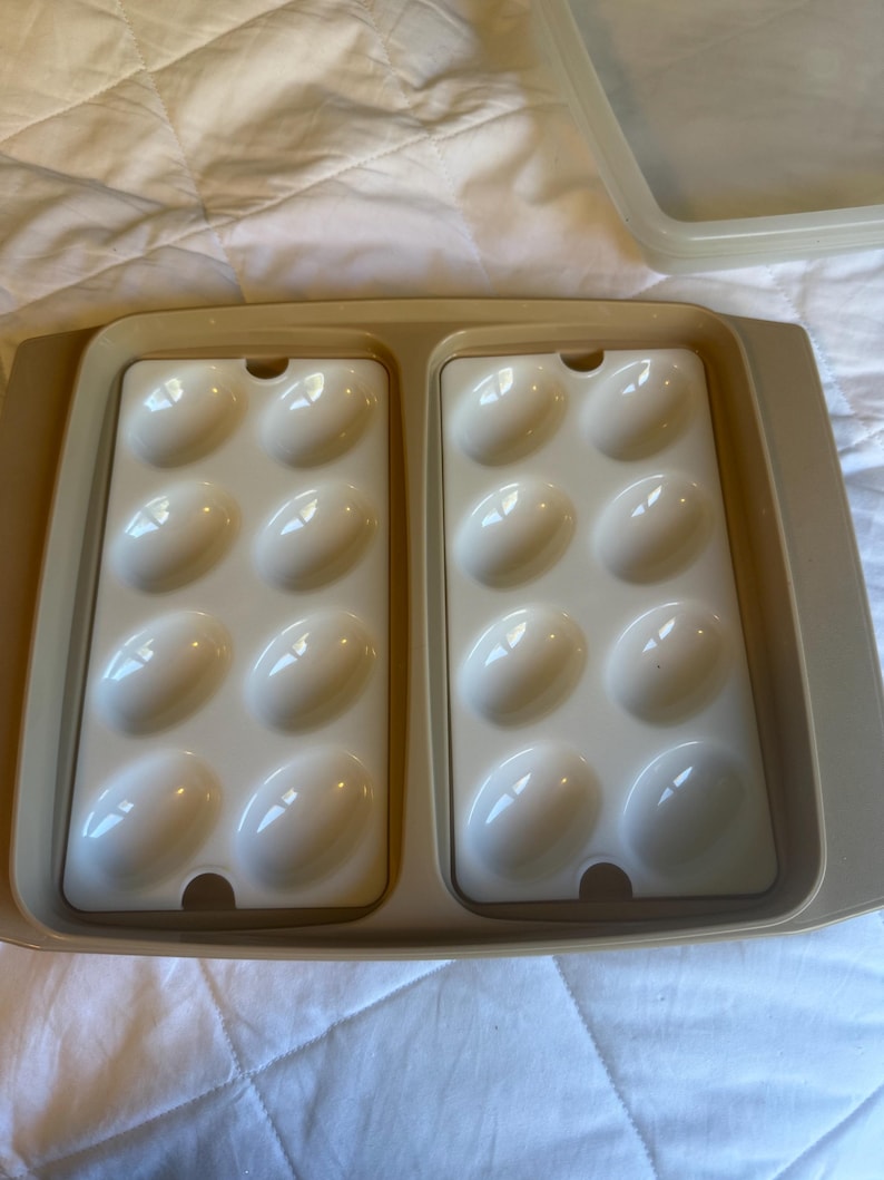 May include: A beige plastic egg container with two trays, each holding six white egg-shaped moulds. The container has a matching lid, designed for storing and transporting eggs. The trays are set inside a larger rectangular base.