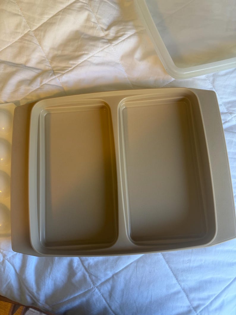 May include: A beige rectangular food storage container with two compartments. The container has a matching lid. The container is made of plastic and is sitting on a white surface.