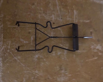 Peut inclure: Un objet en métal noir de forme singulière, avec un triangle central et des lignes courbes. Il comporte un composant cylindrique et une pièce plate argentée qui s'en écarte. L'objet est posé sur une surface texturée brun clair.