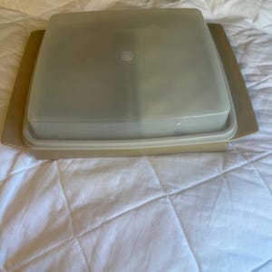 Könnte beinhalten: Ein beigefarbener Tupperware-Behälter mit einem quadratischen, transparenten Deckel. Der Behälter steht auf einer passenden beigefarbenen Basis. Der Deckel hat eine kleine, kreisförmige Vertiefung in der Mitte. Für die Aufbewahrung von Lebensmitteln.