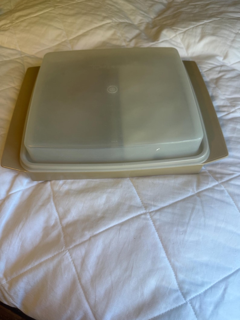 May include: A tan-coloured Tupperware container with a square, clear lid. The container sits on a matching tan base. The lid has a small, circular indentation in the centre. The container is designed for food storage.