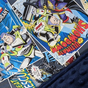 May include: A blanket featuring a comic book panel design with Buzz Lightyear and other Toy Story characters. The blanket has a navy blue, textured border. Comic book panels include the text "BATTLE at GAMMA QUADRANT" and "ULTIMATE SHOWDOWN".