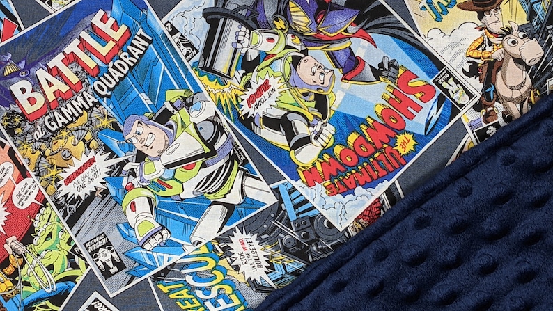 May include: A blanket featuring a comic book panel design with Buzz Lightyear and other Toy Story characters. The blanket has a navy blue, textured border. Comic book panels include the text "BATTLE at GAMMA QUADRANT" and "ULTIMATE SHOWDOWN".