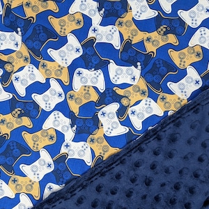 May include: A blue blanket with a pattern of white, gold, and navy blue video game controllers. The blanket has a soft, textured navy blue backing. The blanket is folded diagonally, showing both sides.