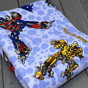 May include: Blue fabric with a repeating pattern of Transformers characters, including Optimus Prime and Bumblebee. The fabric features a blue background with a hexagonal pattern.