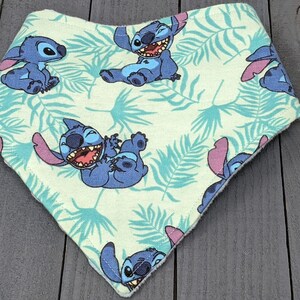 May include: A triangular bandana bib featuring a light green background with a repeating pattern of blue Stitch characters and teal tropical palm leaves. The fabric has a finished stitched edge and a pointed bottom hem in a classic bib shape.