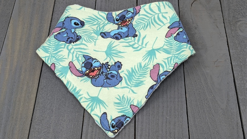May include: A triangular bandana bib featuring a light green background with a repeating pattern of blue Stitch characters and teal tropical palm leaves. The fabric has a finished stitched edge and a pointed bottom hem in a classic bib shape.