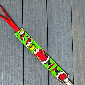 May include: A red and white pacifier clip with a green and white fabric strap featuring a repeating pattern of the Grinch wearing a Santa hat.
