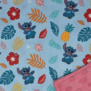 May include: A blue and pink reversible baby blanket featuring a repeating pattern of Stitch from Lilo & Stitch. The pattern includes Stitch dancing in a grass skirt, pineapples, hibiscus flowers, and tropical leaves.