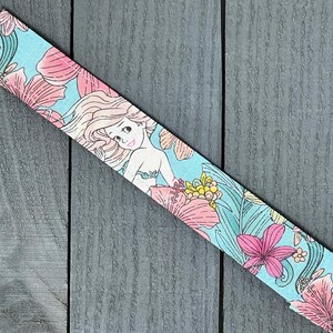 May include: A light blue pacifier clip with a floral pattern featuring a mermaid, pink and yellow flowers, and green leaves. The clip is made of metal and has a white ribbon attached.