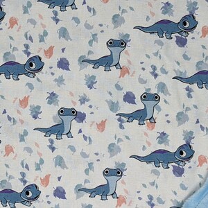 May include: A soft blanket featuring a repeating pattern of blue cartoon salamanders on a white background with scattered small purple and peach-colored leaves. The reverse side of the blanket is a solid light blue fabric with a textured dot pattern.
