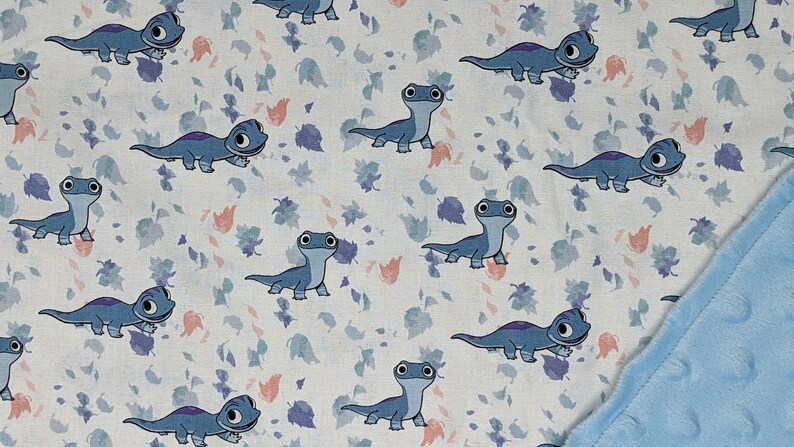 May include: A soft blanket featuring a repeating pattern of blue cartoon salamanders on a white background with scattered small purple and peach-colored leaves. The reverse side of the blanket is a solid light blue fabric with a textured dot pattern.