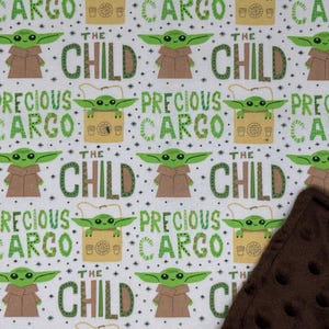 Peut inclure: Tissu blanc avec un motif r&eacute;p&eacute;t&eacute; d'un personnage extraterrestre vert, d'un manteau marron et des mots "THE CHILD" et "PRECIOUS CARGO" en vert. Une couverture marron textur&eacute;e est dans le coin.