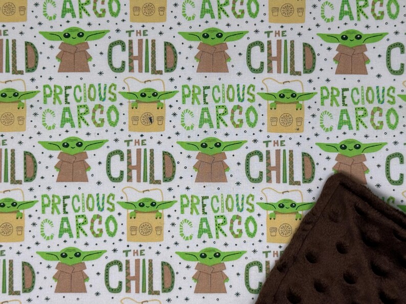 Peut inclure: Tissu blanc avec un motif r&eacute;p&eacute;t&eacute; d'un personnage extraterrestre vert, d'un manteau marron et des mots "THE CHILD" et "PRECIOUS CARGO" en vert. Une couverture marron textur&eacute;e est dans le coin.