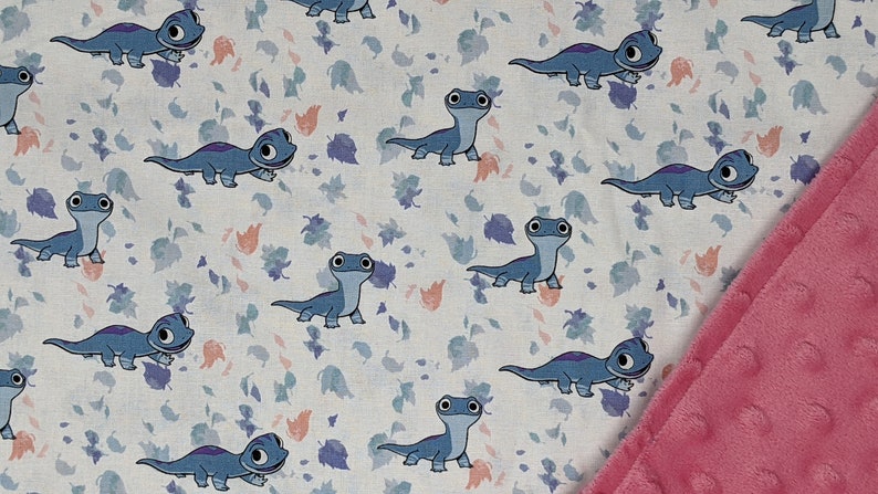 May include: A white fabric with a pattern of blue salamander-like creatures and leaf shapes in blue, purple, and peach. A pink, textured blanket edge is visible on the right side. The design is likely for a baby blanket or similar item.