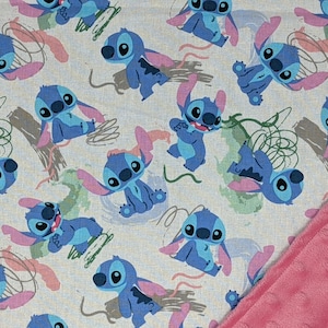 May include: A fabric featuring a repeating pattern of the blue alien character Stitch in various poses, interspersed with green and brown abstract shapes on a light background, with a corner of textured pink dimpled fabric folded over.