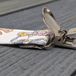 May include: A white fabric strap with a cartoon print of animals and people. The strap has a silver metal clip with a gold screw.