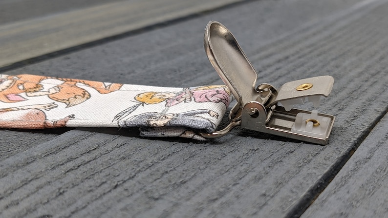 May include: A white fabric strap with a cartoon print of animals and people. The strap has a silver metal clip with a gold screw.