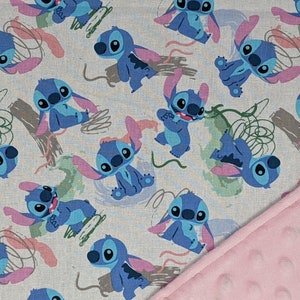 May include: A fabric blanket featuring a repeating pattern of the blue character Stitch from Lilo and Stitch on a light gray background, with abstract green and pink brush strokes, paired with a solid light pink textured dimple dot fabric backing.
