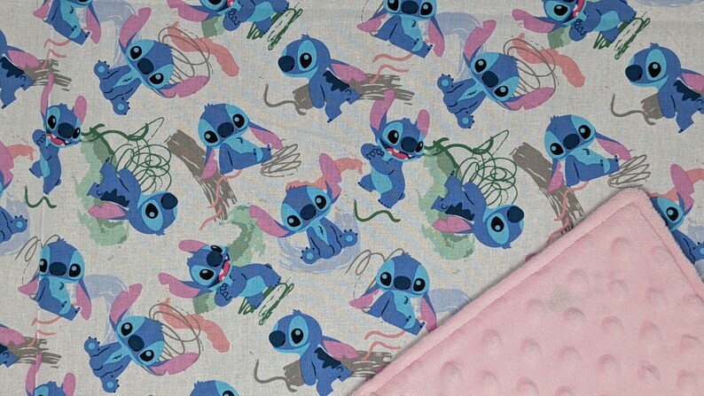 May include: A fabric blanket featuring a repeating pattern of the blue character Stitch from Lilo and Stitch on a light gray background, with abstract green and pink brush strokes, paired with a solid light pink textured dimple dot fabric backing.