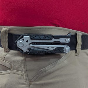 May include: A multi-tool clipped to a black belt worn with khaki pants and a red shirt. The multi-tool is silver and black, with various tools visible. The belt is secured around the waist.