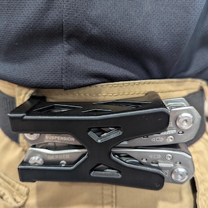 May include: A black multi-tool holder with two silver multi-tools clipped to a tan belt. The multi-tools are labeled "GERBER" and "SUSPENSION". The holder is made of black metal and is attached to a tan belt.