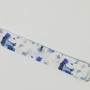 May include: A fabric pacifier clip featuring a white background with a repeating pattern of blue cartoon hammerhead sharks and scattered purple and blue leaf-like shapes. The strap includes a light blue ribbon loop and a silver metal clip.