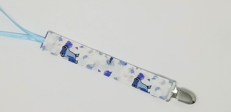 May include: A fabric pacifier clip featuring a white background with a repeating pattern of blue cartoon hammerhead sharks and scattered purple and blue leaf-like shapes. The strap includes a light blue ribbon loop and a silver metal clip.