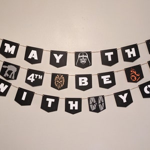 May include: Black and white banner with the text "May the 4th be with you" and Star Wars characters. The banner is strung with twine and has a white background.