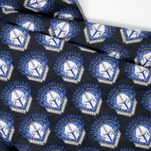 K&ouml;nnte beinhalten: Schwarzer Stoff mit einem sich wiederholenden Muster, das ein Mandalorianer-Helm-Design zeigt. Das Design enth&auml;lt die W&ouml;rter "Mandalorian Warrior Hunter Bounty" in Wei&szlig; und Grau mit blauen Akzenten.
