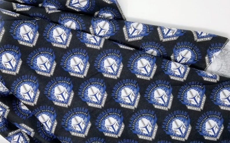 K&ouml;nnte beinhalten: Schwarzer Stoff mit einem sich wiederholenden Muster, das ein Mandalorianer-Helm-Design zeigt. Das Design enth&auml;lt die W&ouml;rter "Mandalorian Warrior Hunter Bounty" in Wei&szlig; und Grau mit blauen Akzenten.