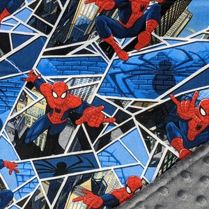 K&ouml;nnte beinhalten: Ein gemusterter Stoff mit Spider-Man in verschiedenen Actionposen. Das Design beinhaltet einen blau-wei&szlig;en geometrischen Hintergrund mit Stadtlandschaften und Spinnennetzdetails. Ein grauer, geriffelter Stoff ist unten rechts sichtbar.