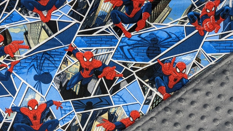 K&ouml;nnte beinhalten: Ein gemusterter Stoff mit Spider-Man in verschiedenen Actionposen. Das Design beinhaltet einen blau-wei&szlig;en geometrischen Hintergrund mit Stadtlandschaften und Spinnennetzdetails. Ein grauer, geriffelter Stoff ist unten rechts sichtbar.