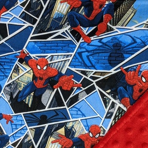 K&ouml;nnte beinhalten: Eine Decke mit einem sich wiederholenden Muster von Spider-Man in Actionposen vor Stadtlandschaften und Spinnennetzen. Die Decke hat einen roten, strukturierten Rand.