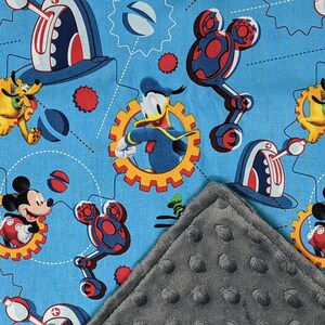 Puede incluir: Una manta para beb&eacute; con un fondo azul claro y un patr&oacute;n repetido de Mickey Mouse, Donald Duck, Pluto y Goofy. El dise&ntilde;o incluye engranajes y elementos mec&aacute;nicos en rojo, blanco y azul. Se ve una esquina gris texturizada.