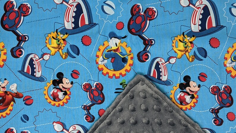 Puede incluir: Una manta para beb&eacute; con un fondo azul claro y un patr&oacute;n repetido de Mickey Mouse, Donald Duck, Pluto y Goofy. El dise&ntilde;o incluye engranajes y elementos mec&aacute;nicos en rojo, blanco y azul. Se ve una esquina gris texturizada.